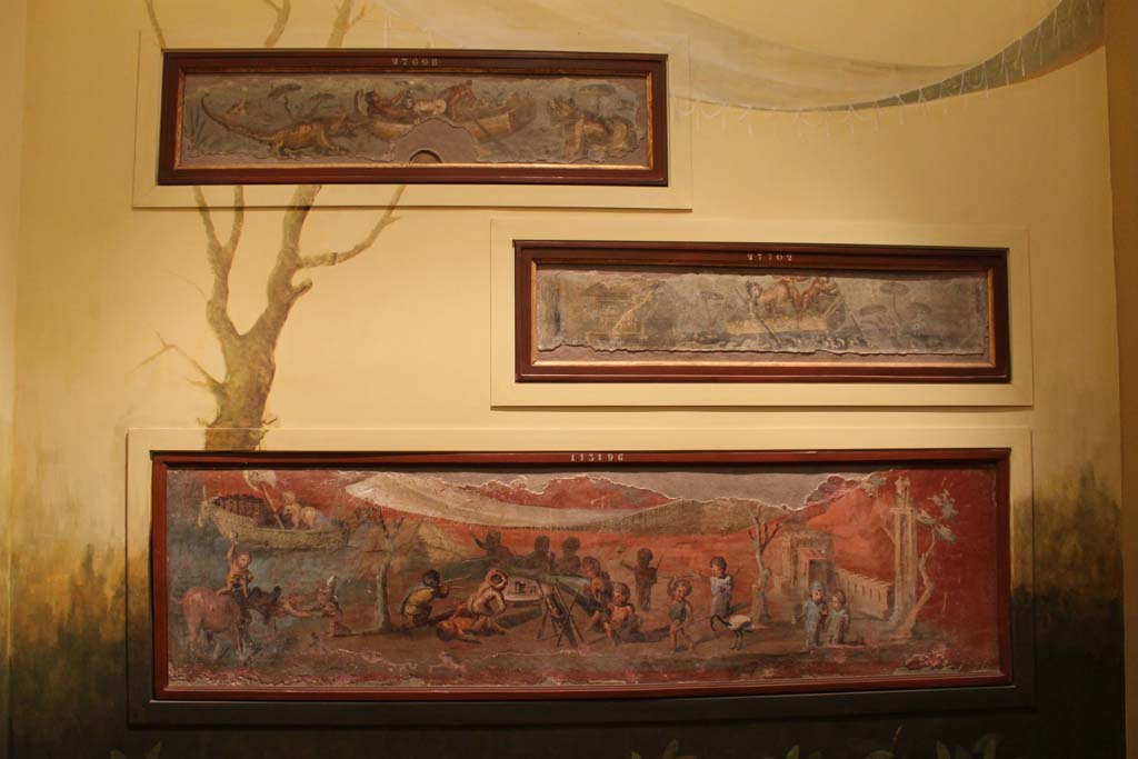 VIII.5.24 Pompeii. March 2014. Display of painting on a wall in Naples Archaeological Museum.
The lowest painting is a Nile banquet scene with pygmies. From a low wall connecting peristyle columns in VIII.5.24.  
The upper paintings are from VII.2.25.
Foto Annette Haug, ERC Grant 681269 DÉCOR.
