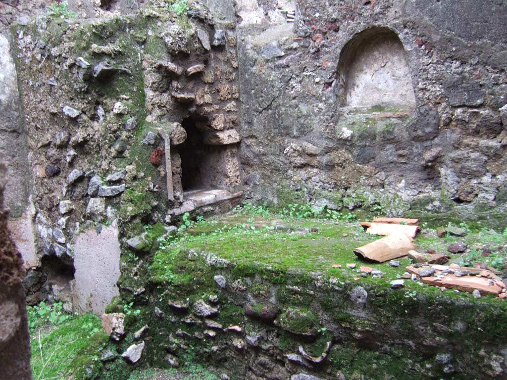 V.1.11 Pompeii. December 2005. Kitchen, hearth and niche, looking east.
According to Boyce, the arched niche on the east wall above the hearth had two holes in its floor for the bases of statuettes.
Only a faint trace of the original lararium painting on the wall beside the niche remained (when he wrote in 1937).
See Boyce G. K., 1937. Corpus of the Lararia of Pompeii. Rome: MAAR 14. (p.32, no.75) 
