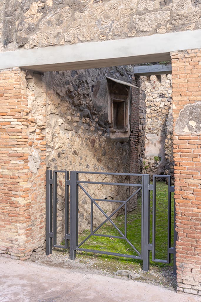 V.1.28 Pompeii. January 2024. 
Looking towards north side of entrance fauces/corridor, with niche. Photo courtesy of Johannes Eber.
According to Boyce, the square niche was surrounded by an elaborate aedicula façade of stucco.
In the floor of the niche were holes for the bases of statuettes.
The niche and façade were brightly coloured, the rear wall was white bordered with red and having delicate painted green plants on it.
The pilasters of the façade were yellow, their capitals red, white and blue.
The bands of relief that ran around the architrave and the cornices were also painted in these colours.
On the wall below the niche was a painted band of yellow.
See Boyce G. K., 1937. Corpus of the Lararia of Pompeii. Rome: MAAR 14. (p.33, no.81, and Pl.1,5) 
Described by Giacobello as a Pseudo-aedicula lararium.
See Giacobello, F., 2008. Larari Pompeiani: Iconografia e culto dei Lari in ambito domestico. Milano: LED Edizioni, (p.231, no.F4)

