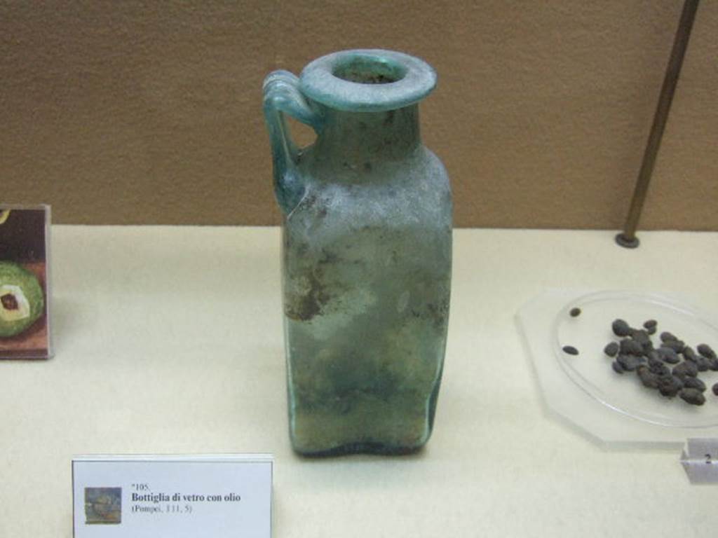 I.11.5 Pompeii. Square glass bottle with round neck, flared rim and with handle. It was used for oil. Inside was found a yellow coloured solid. Tests showed this was oil.  Now in Boscoreale Antiquarium. SAP Inventory number 12821. See Cibi e Sapori a Pompei e Dintorni, exhibition catalogue from Boscoreale Antiquarium, 2005, (p. 89, No. 105).