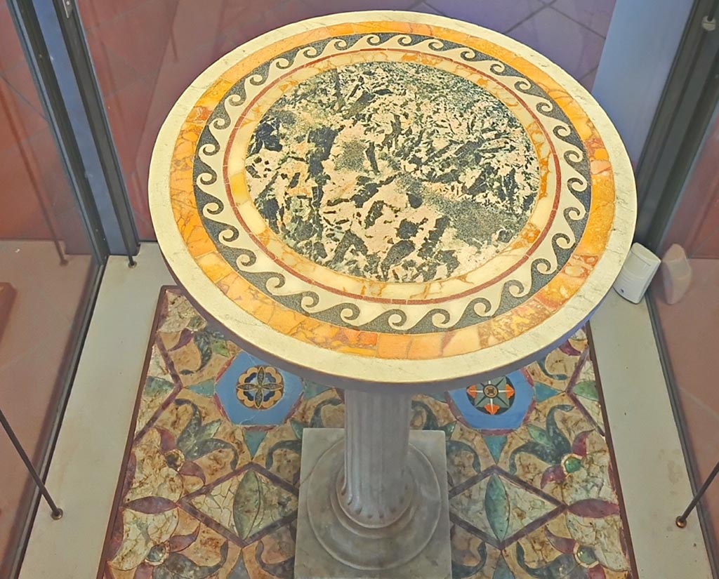 V.1.18 Pompeii. September 2024. Marble table. Photo courtesy of Giuseppe Ciaramella.
According to the information card –
Inventory number – 120530.
The table consists of a white marble top, decorated by polychrome inlay with a green marble tondo surrounded by a black marble wave pattern, placed on a small column which is not an original feature.
(Second–third quarter of the Ist century AD).
(Tavolo marmoreo. Tavolo composto da un piano in marmo bianco decorato da commessi policromi, con tondo in marmo verde circondato da un motivo a onda corrente in marmo nero, su colonnina di sostegno non pertinente. Secondo – terzo quarto del I secolo d.C.)

