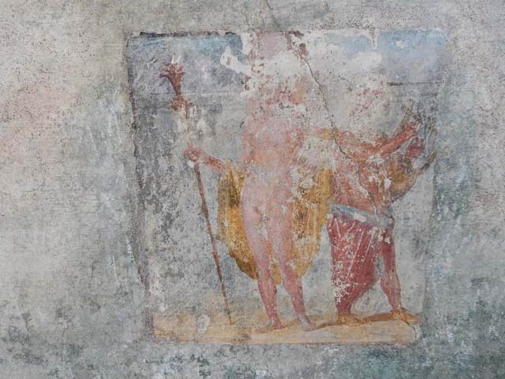 V.4.a Pompeii. May 2015. Room ‘s’, wall painting of Dionysus (Bacchus) accompanied by Silenus playing the lyre. 
From the east wall of the summer triclinium. Photo courtesy of Buzz Ferebee.
According to Kuivalainen, who describes the painting as –
“A composition of two standing figures on yellowish terrain against a simple architectural background. 
An effeminate youth with fair complexion stands frontally, with his weight on his right foot; in his right hand he holds a thyrsus with a bunch of leaves and a torch-like decoration on the top; ……..”
Kuivalainen then comments –
“Though the pose is customary to practically naked Bacchus leaning on a playing Silenus, the figure would possibly be a hermaphrodite, not Bacchus. This painting had a parallel in room ‘t’ of the House of Caecilius Iuncundus (V.1.26, our room 22). The torch-like thyrsus would be a rarity to Bacchus. It is more common for hermaphrodites and divinities of light.”
See Kuivalainen, I., 2021. The Portrayal of Pompeian Bacchus. Commentationes Humanarum Litterarum 140. Helsinki: Finnish Society of Sciences and Letters, (F14, p.170).

