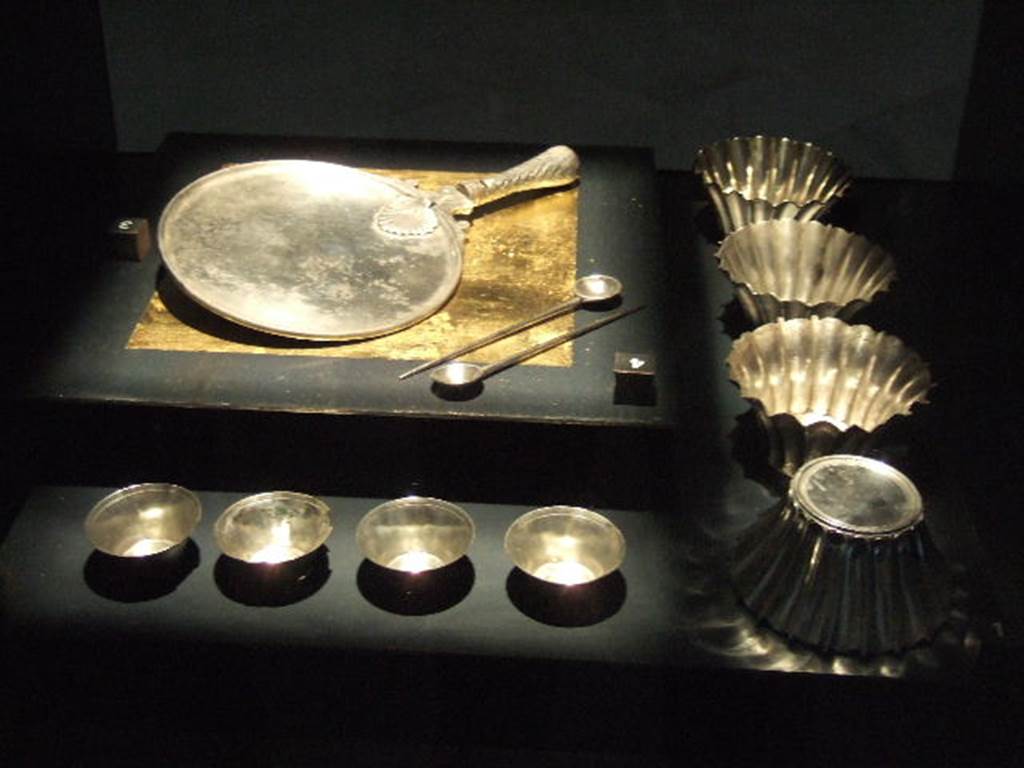 Silver found at VI.7.19.  Now in Naples Archaeological Museum
Four lanceolate pattern silver formae or moulds (on the right side). Inventory numbers 25552, 25553, 25554 and 25555.
Three of the moulds (25552, 25553, 25554) have inscriptions on them. These give the names of Helvius Amandus and L. Herrenius Rusticus.
According to Epigraphik-Datenbank Clauss/Slaby (See www.manfredclauss.de) these read

Helvi Amandi p(ondo) I(librae) s(emissem) |(unciarum) |(semunciae) III / [3] Herenni Rustici p(ondo) I(librae) s(emissem) |(unciarum) [3]II       [CIL X 8071,09a]
Helvi Amandi p(ondo) I(librae) s(emissem) |(unciarum) III / L(uci) Herenni Rustici p(ondo) I s(emissem) |(unciarum) II        [CIL X 8071,09b]
Helvi Amandi p(ondo) I(librae) s(emissem) IV(unciarum) s(emunciae) III        [CIL X 8071,09c]

Large silver mirror with a diameter of 18cm (top left). Inventory Number 25718.
Two silver spoons (centre), part of a group of five. Inventory numbers 25428 and 25429.
Four small round silver moulds in the form of a truncated cone (front left). Inventory numbers 25329, 25330, 25331 and 25332.
See Guzzo, P. (A cura di), 2006. Argenti a Pompei. Milano, Electa. (p. 168-179).
