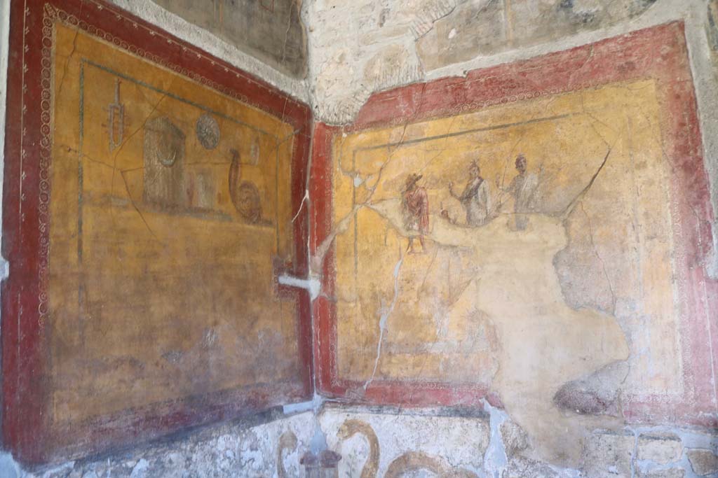 VI.16.7 Pompeii. May 2016. Information card. Photo courtesy of Buzz Ferebee.
“The second lararium, painted in the south-eastern corner of the peristyle, was instead dedicated to the Egyptian deities: portrayed on a tall base with two serpents, converging towards two altars upon which eggs have been placed as an offering, is the family of Isis, Serapides and Harpocrates. With them is Anubis, the god of the dead who has a canine head, holding a caduceus symbolizing his assimilation with Mercury.  Next to them are the objects of the cult of Isis, which also express a domestic type of devotional practice: these include a sistrum, a cist, a silver situla in the shape of a human breast to contain holy milk, and a patera also made of silver. 
Watching over them is Uraeus, the sacred cobra.”

