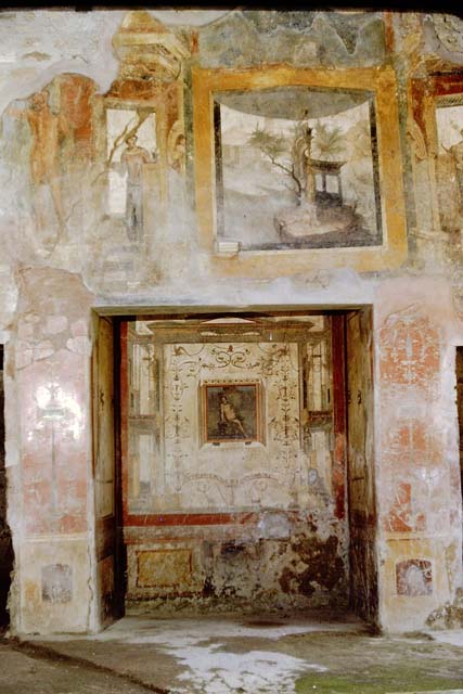 VI.16.15 Pompeii. December 2006. North wall of atrium with lararium between doorways to room I (left) and rooms K to M (right).
According to Boyce, the space between the two doorways on the north side of the atrium was painted with white stucco, bordered with red stripes.
This formed the large panel for the lararium painting.
A square niche was in the upper right-hand corner, on the rear wall of which was painted the figure of the Genius.
Hanging down on each side of him was a garland, on the side walls were painted red flowers with green leaves.
Of the two Lares originally painted on the white background of the wall, one on each side of the niche, only the lower part of the one on the left remained.
Below the figure of the Lar was painted a tall cylindrical altar, furnished with two eggs, a pine cone and fruit.
On each side was a huge yellow serpent, each coiled serpent about two metres in length.
The one on the left had a red crest and beard. The one on the right had neither.
In the background were rather large plants bearing fruit.
Over the surface of this picture had been spread a thin wash.
It would appear that the lararium belonged to an earlier decoration of the house and was to be replaced by a new one to be painted on the thin wash.
See Notizie degli Scavi di Antichità, 1908, p. 67.
See Boyce G. K., 1937. Corpus of the Lararia of Pompeii. Rome: MAAR 14. (p.58, no.224, Pl.16,1) 
See Fröhlich, T., 1991. Lararien und Fassadenbilder in den Vesuvstädten. Mainz: von Zabern. (L75, picture. 39,2)
See Giacobello, F., 2008. Larari Pompeiani: Iconografia e culto dei Lari in ambito domestico. Milano: LED Edizioni. (p.185)
