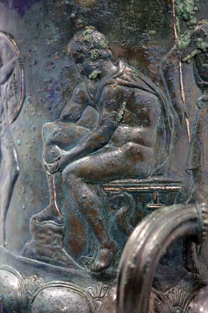IX.13.1-3 Pompeii. February 2021. 
Bronze krater with scenes in relief, found in triclinium, on display in Antiquarium, VIII.1.4.
According to the description card in the Antiquarium -
The bronze krater was decorated with eight armed male figures, six standing and two sitting. 
Detail of one of the seated figures tying his shoelaces.
Photo courtesy of Fabien Bièvre-Perrin (CC BY-NC-SA).
