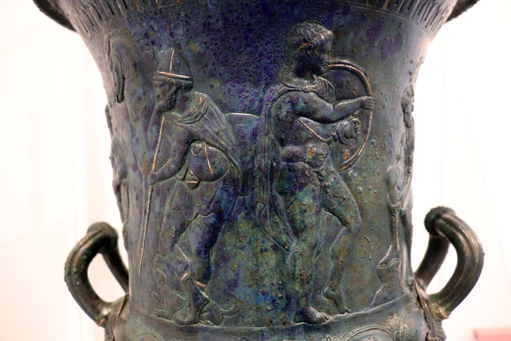 IX.13.1-3 Pompeii. February 2021. 
Detail from bronze krater with scenes in relief, found in triclinium, on display in Antiquarium, VIII.1.4.
Standing in front of the seated figure tying his shoelaces are a young man holding a shield, with a bearded figure holding a staff behind.
Photo courtesy of Fabien Bièvre-Perrin (CC BY-NC-SA).
