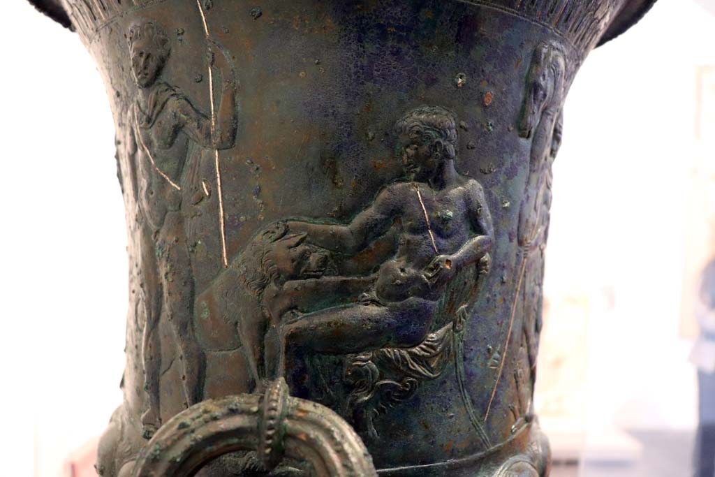 IX.13.1-3 Pompeii. February 2021. 
Bronze krater with scenes in relief, found in triclinium, on display in Antiquarium, VIII.1.4.
Detail of the second sitting figure, young and naked, caressing an animal with one hand and holding a sword by the hilt in the other.
