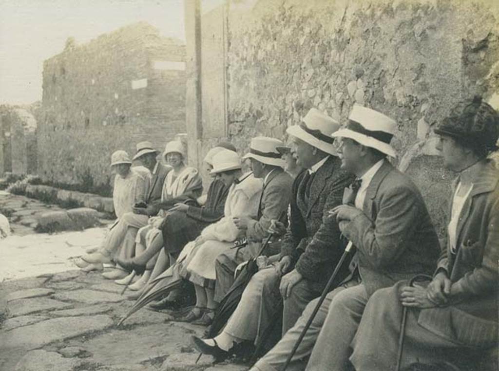 Vicolo Storto, Pompeii. 5th June 1925. Tourists having a rest sitting on the high pavement on the west side of the vicolo. On the left is the junction of Vicolo Storto, with the Via degli Augustali, and on the opposite side of the roadway is the Vicolo di Eumachia. Photo courtesy of Rick Bauer.
