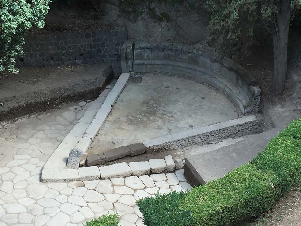 NGH Pompeii. 2015. Schola tomb after clearance showing previously covered tomb and basalt block roads. 
The continuation of ancient Via di Nola into unexcavated area is on the left.
The area immediately outside of Porta Nola (the Nolan Gate) was first cleared in 1907–8. 
Since then, soil had once again accumulated opposite the gate, re-burying the tomb. 
In 2015 the Porta Nola Project has cleared the basalt block roads and brought the monument back to light.
Excavation inside the monument in front of the benches revealed a thick layer of floor preparation for the pavement which has since disappeared.
See BSR Pompeii Project Porta Nola Necropolis 2015 Excavations
