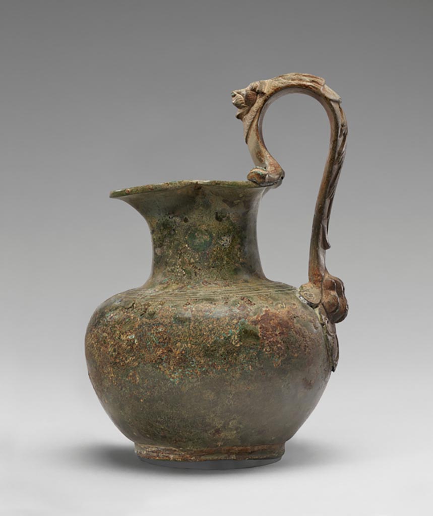 Discovered in a villa in Boscoreale. Bronze oinochoe (pitcher) with lion handle.
Digital image courtesy of the Getty's Open Content Program. Now in the Getty Museum, inventory number 72.AC.146.
The shoulder of this otherwise plain oinochoe (pitcher) is decorated with two pairs of incised bands. Parts of the high, arched handle, however, take the form of a lion. A lion’s head in relief appears above the vessel’s mouth. The rotellae, small protrusions on either side of the handle where it joins the rim, are fashioned as the stylized forelegs of the feline. At the base of the handle is a lion’s paw. Floral motifs run along the outer face of the handle.
Vessels of this type were used in the home for religious rituals and to hold water for handwashing during banquets. Slaves would pour water from an oinochoe over the hands of participants, catching the cascade in a patera (shallow bowl) held underneath. This oinochoe was discovered in a villa at Boscoreale or in the vicinity, although the exact findspot is unknown. 